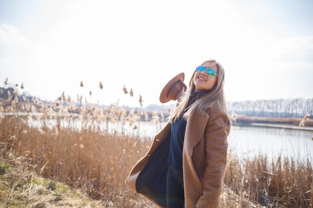 Una niña con un abrigo marrón, sombrero y gafas camina en un parque con un lago bajo el sol brillante. Se regocija en la vida y sonríe. El comienzo de la primavera