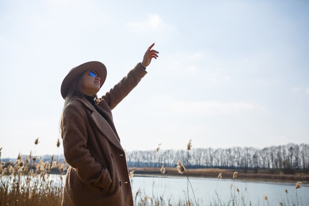 Una niña con un abrigo marrón, sombrero y gafas camina en un parque con un lago bajo el sol brillante. Se regocija en la vida y sonríe. El comienzo de la primavera