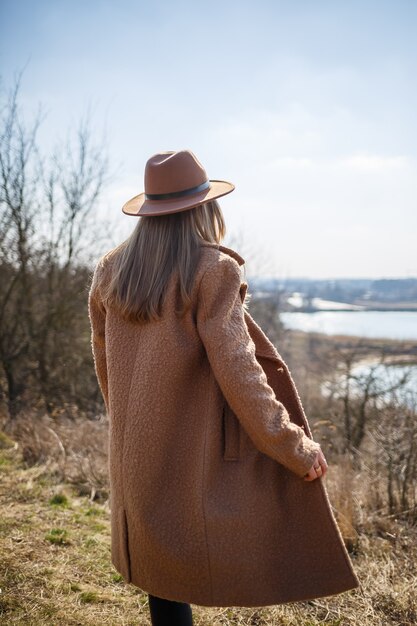 Una niña con un abrigo marrón, sombrero y gafas camina en el parque con un lago bajo el sol brillante.