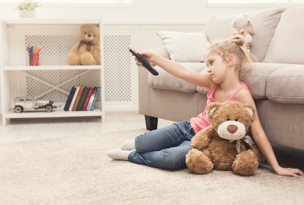 Niña aburrida viendo la televisión en casa. Niña sentada en la alfombra del piso con su amigo de juguete, el oso de peluche, sosteniendo el control remoto y cambiando de canal, nada interesante que ver, copie el espacio