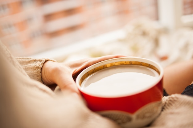 La niña en un acogedor suéter de punto tomando café de una taza roja