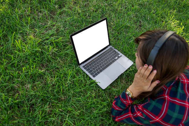 Una niña acostada en un prado usa su computadora portátil y escucha música en una pantalla en blanco con una maqueta