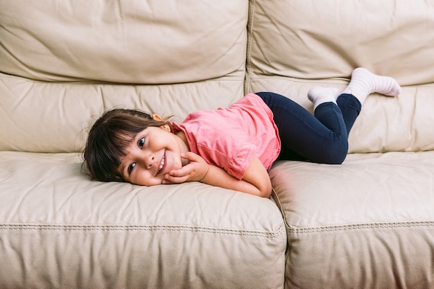 Una niña, acostada y sonriendo en un sofá color crema con una camiseta rosa y pantalón azul