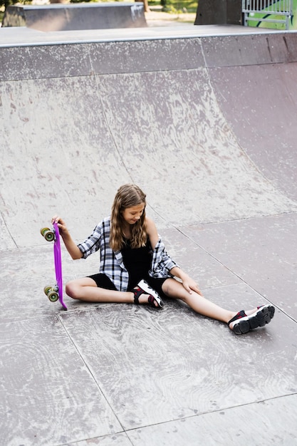 Niña activa después de caer de un tablero de penny lesionado sentado y mirando un hematoma en la rampa deportiva en el patio de recreo del parque de patinaje