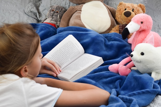 niña se acuesta en la cama con un libro y juguetes