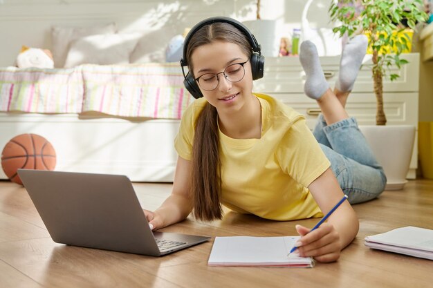 Foto niña adolescente con auriculares usando una computadora portátil tendida en el suelo escribiendo en un cuaderno