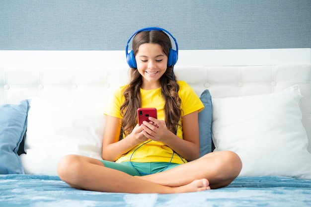 Niña adolescente con auriculares viendo videos en un teléfono inteligente sentada en la cama en su habitación Estudiante adolescente sonriente estudiando en línea con elearning de teléfono móvil