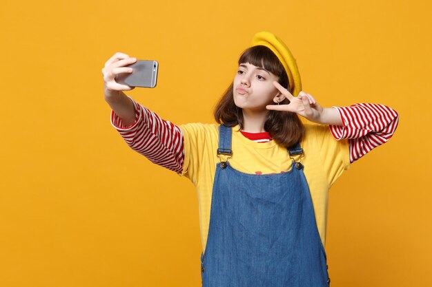 Niña adolescente con boina francesa, vestido de mezclilla haciendo selfie en el teléfono móvil, mostrando el signo de la victoria soplando labios aislados en un fondo amarillo. Concepto de estilo de vida de las emociones de las personas. Simulacros de espacio de copia.