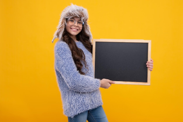 Niña adolescente en cálido sombrero de otoño y suéter sosteniendo pizarra aislada sobre un fondo amarillo Maqueta de espacio de copia de venta de escuela de otoño Niña feliz cara emociones positivas y sonrientes