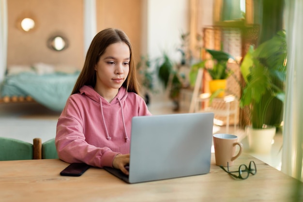 Niña adolescente enfocada que estudia en línea usando una computadora portátil sentada en el escritorio en casa con una lección en línea escribiendo en el teclado