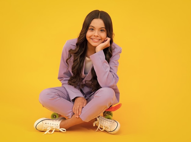 Niña adolescente hermosa y de moda con traje moderno posando con una patineta aislada de fondo amarillo