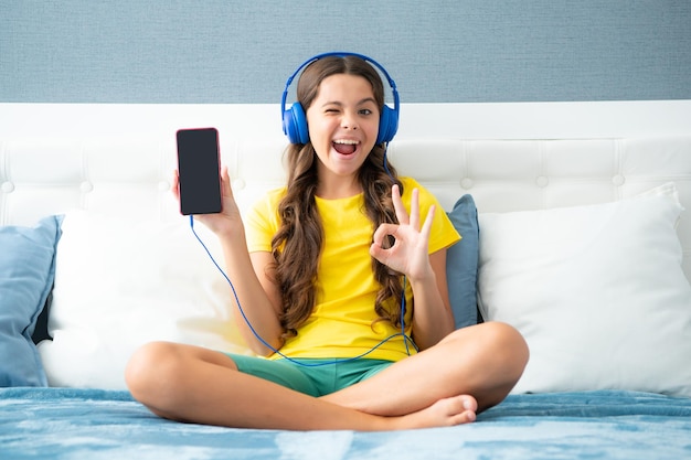 Niña adolescente mostrando un teléfono con pantalla usando audífonos escuchando música en una sesión de teléfono inteligente