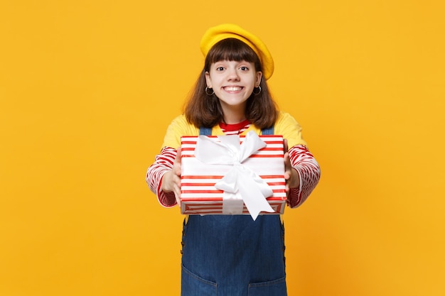 Niña adolescente sonriente con boina francesa, vestido de mezclilla con caja de regalo a rayas rojas con cinta de regalo aislada en el fondo de la pared amarilla. Emociones sinceras de la gente, concepto de vacaciones de cumpleaños de estilo de vida.