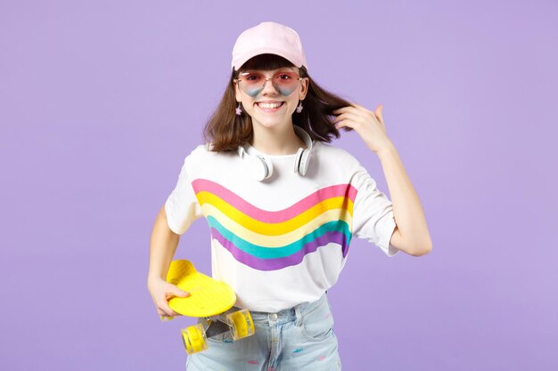 Niña adolescente sonriente con ropa vívida, anteojos de corazón con auriculares sosteniendo patineta amarilla aislada en el fondo de la pared pastel violeta. Gente emociones sinceras concepto de estilo de vida. Simulacros de espacio de copia.