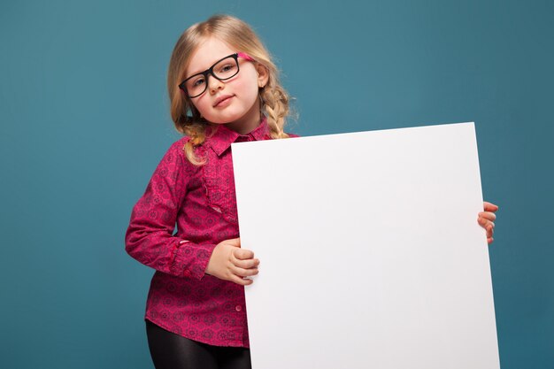 La niña adorable en camisa rosada, pantalones negros y vidrios sostiene el cartel vacío