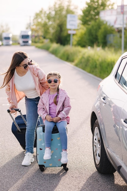 Una niña adorable se sienta en la maleta de su hijo y espera el viaje con su madre. Un auto está estacionado a un lado.