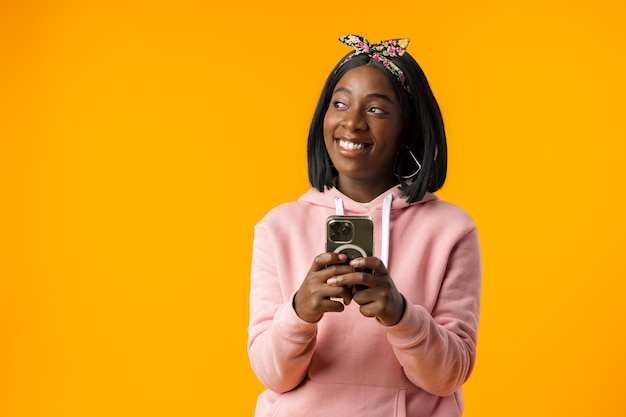 Niña africana feliz usando su teléfono inteligente con fondo amarillo