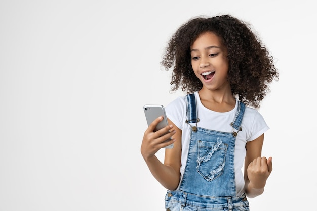 Niña africana joven feliz usando teléfono móvil hacer gesto de ganador posando aislado