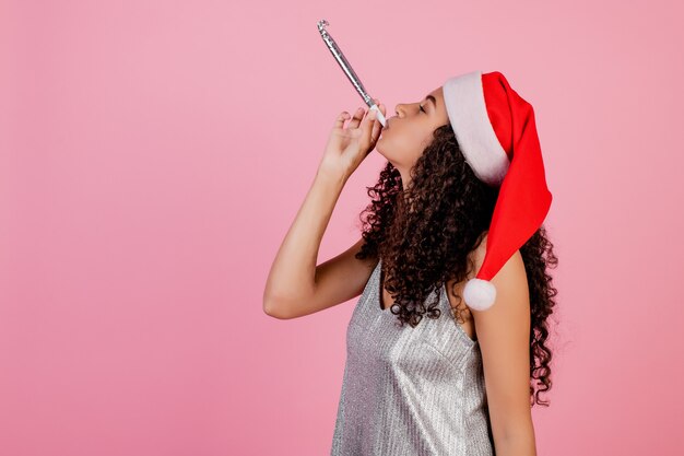 Foto niña africana que sopla silbato de navidad con sombrero de navidad aislado en rosa