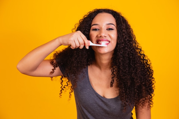 Niña afro cepillarse los dientes sobre fondo amarillo. Mujer afro cepillándose los dientes. Concepto de salud bucal