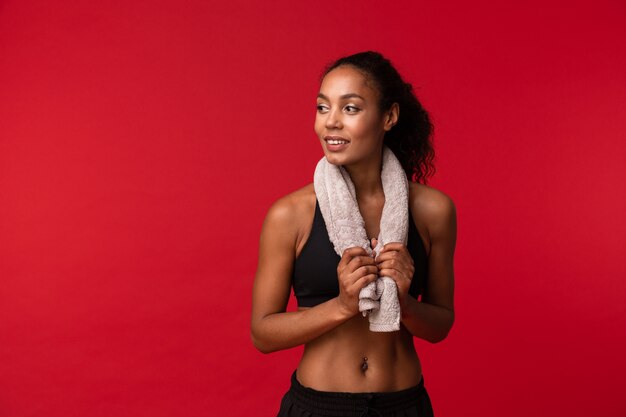 Niña afroamericana de 20 años en ropa deportiva negra con una toalla sobre su cuello de pie, aislado sobre una pared roja