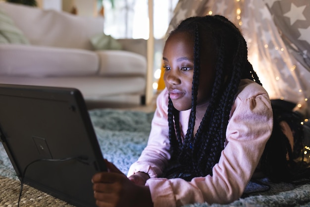 Niña afroamericana acostada sobre el estómago usando una tableta en casa luces de cuerda de tienda de estrellas copia el espacio