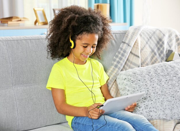 Niña afroamericana con auriculares y tablet en el sofá de la habitación