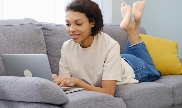 Foto niña afroamericana confiada que usa una computadora portátil en un teléfono inteligente sentada en un sofá en el espacio de copia de casa