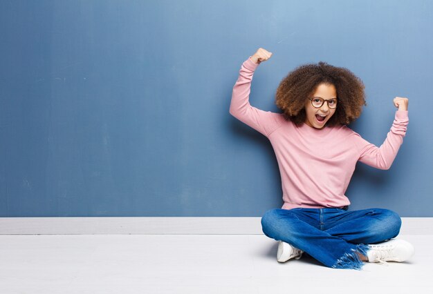 Niña afroamericana gritando triunfante, luciendo emocionada, feliz y sorprendida ganadora, celebrando sentada en el suelo