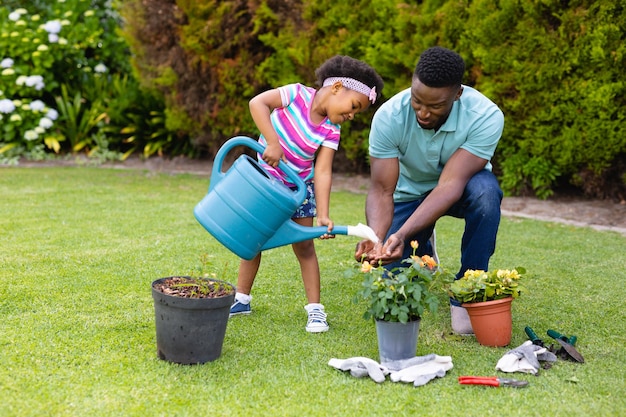 Niña afroamericana y padre adulto medio con regadera jardinería en el patio trasero