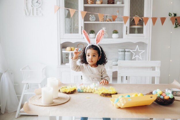 Niña afroamericana pinta huevos de Pascua en casa en la cocina felices pascuas