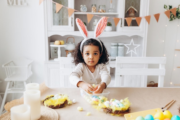 Niña afroamericana pinta huevos de Pascua en casa en la cocina felices pascuas