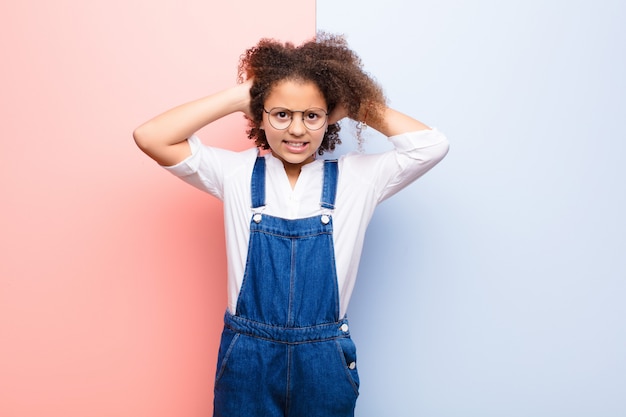 Niña afroamericana que se siente estresada, preocupada, ansiosa o asustada, con las manos en la cabeza, en pánico por el error contra la pared plana