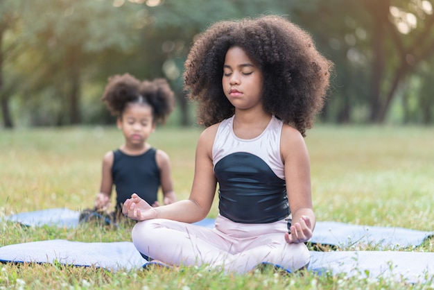 Niña afroamericana sentada en la estera del rollo practicando meditar yoga en el parque al aire libre