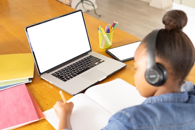 Niña afroamericana sentada en la mesa usando una computadora portátil y una tableta con espacio para copiar lecciones en línea