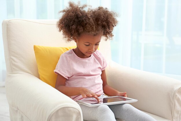 Niña afroamericana sentada en un sillón con tableta en la habitación