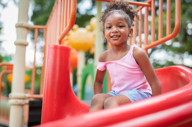 La niña afroamericana sonriente se divierte jugando en el control deslizante en el patio de recreo en el parque