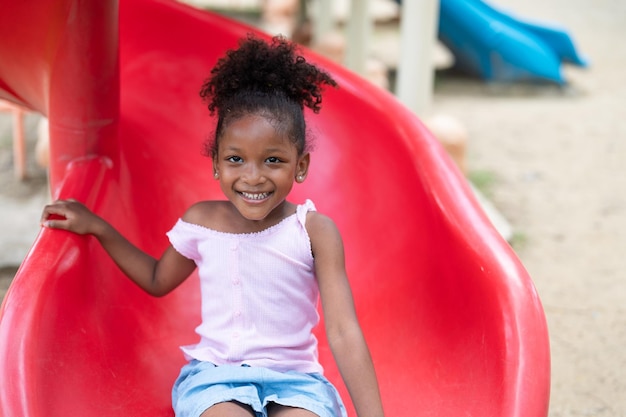 La niña afroamericana sonriente se divierte jugando en el control deslizante en el patio de recreo en el parque