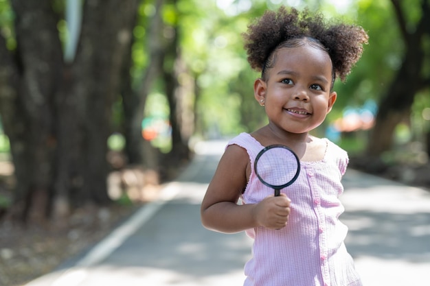 Niña afroamericana sonriente se divierte sosteniendo una lupa para explorar en el árbol en el parque