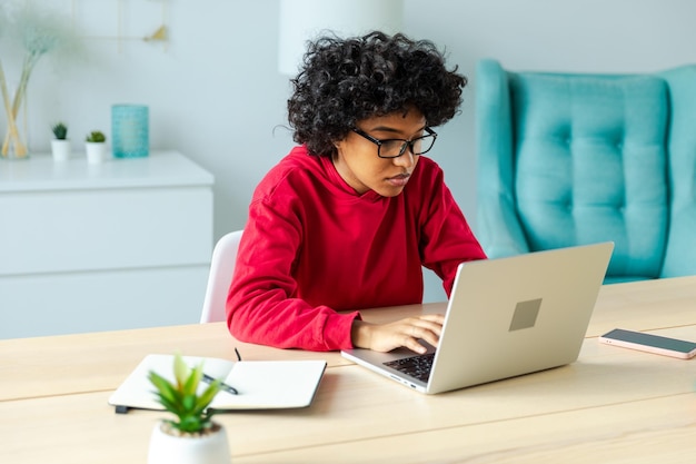 Niña afroamericana usando una computadora portátil en la oficina de casa mirando la pantalla escribiendo charlando leyendo escribiendo