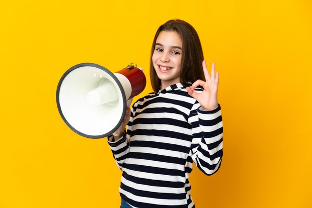 Niña aislada en la pared amarilla sosteniendo un megáfono y mostrando el signo de ok con los dedos