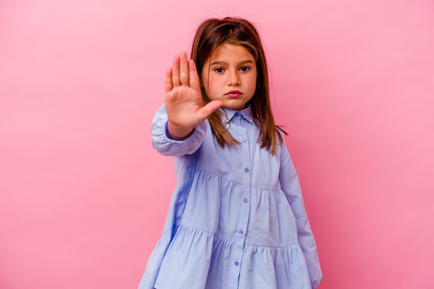 Niña aislada en la pared de color rosa de pie con la mano extendida mostrando la señal de stop, impidiéndole