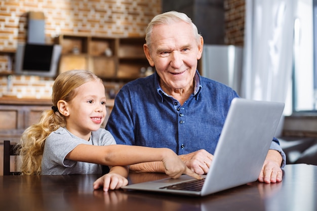 Niña alegre apuntando la pantalla mientras usa la computadora portátil con su abuelo