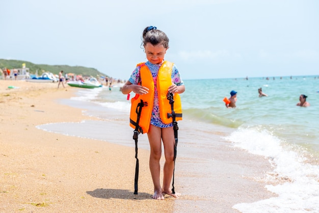 Una niña alegre con un chaleco salvavidas se para en la playa y mira la seguridad del agua de las olas