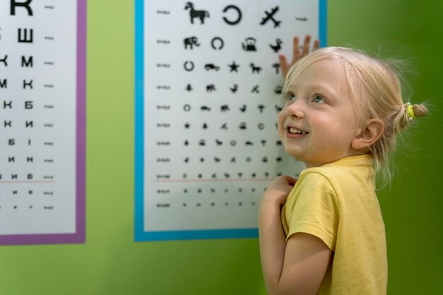 Niña alegre en la cita con el optometrista cerca del cartel para revisar su vista Optometrista pediátrico