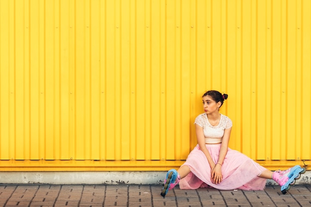 Una niña alegre come helado y rueda sobre rodillos en verano contra una pared amarilla.