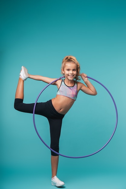 Niña alegre de deportes haciendo ejercicios con un hula hoop aislado sobre pared azul