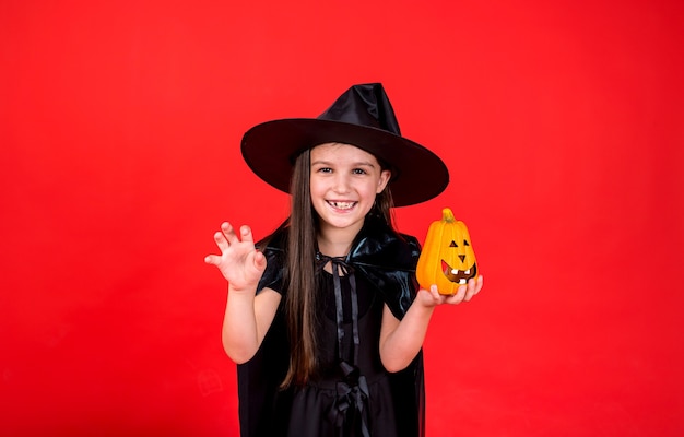 Una niña alegre en un disfraz de bruja sostiene una calabaza sobre un fondo rojo con una copia del espacio