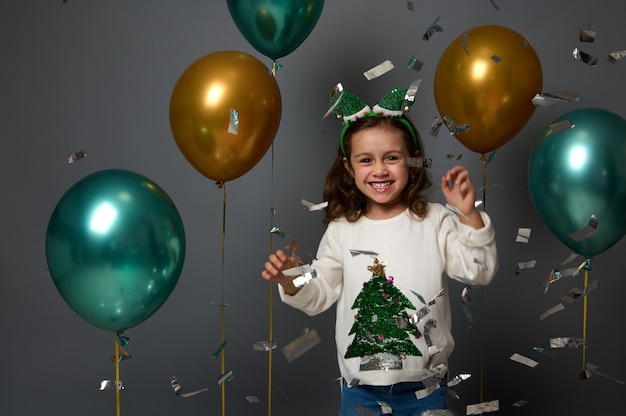 Niña alegre en elf hoop divirtiéndose en la fiesta de año nuevo lanzando confeti de lentejuelas, regocijándose contra el fondo gris con globos de aire inflados de color verde dorado brillante. Concepto de Navidad, espacio publicitario de copia