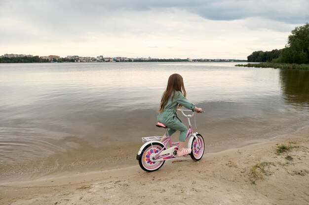 Niña alegre feliz con su bicicleta en la banda del río en la naturaleza